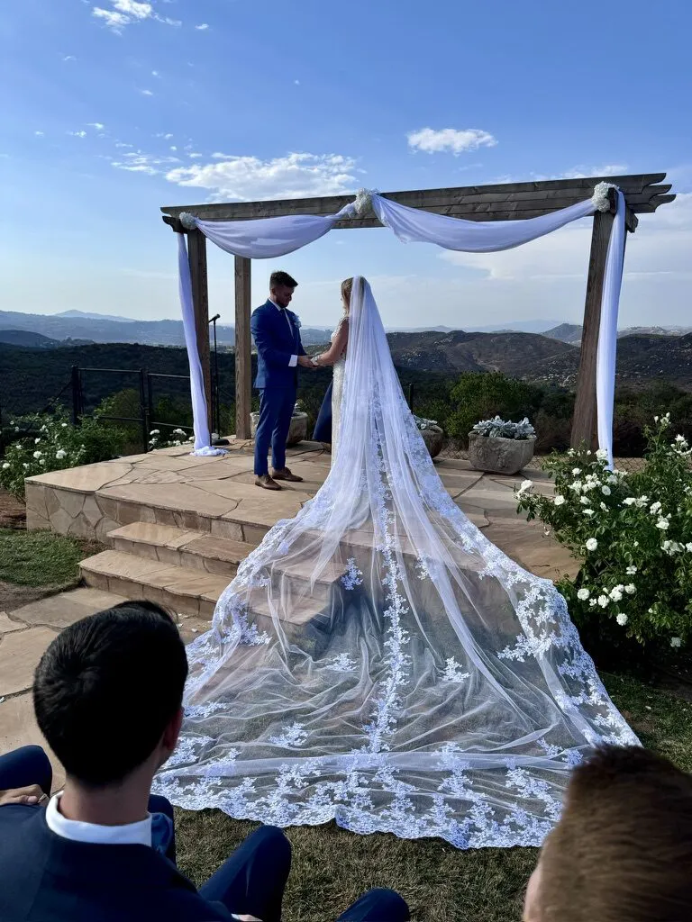 Couple exchanging vows at the draped pergola, the bride's cathedral veil flowing down the stone steps